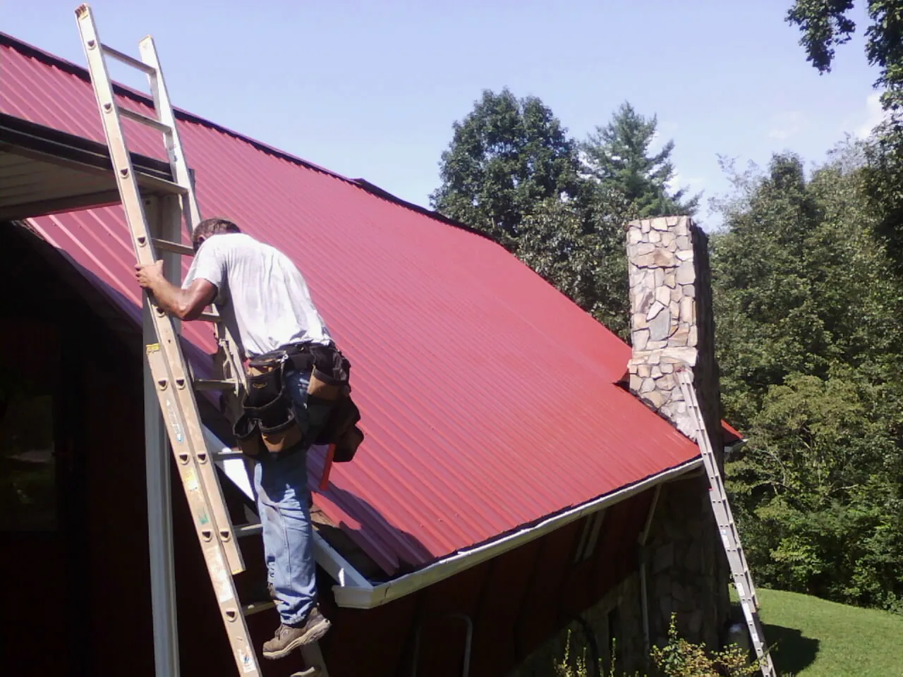 Roofer on a ladder beside a red metal roof and stone chimney — Soffit & Fascia Repair by Roofing Brothers in Edmeston